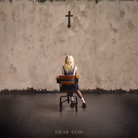 A blonde woman in a navy tartan school pinafore sits at a wooden school desk facing a bare plaster wall, with a black cross mounted above her that tapers into a downward arrow.