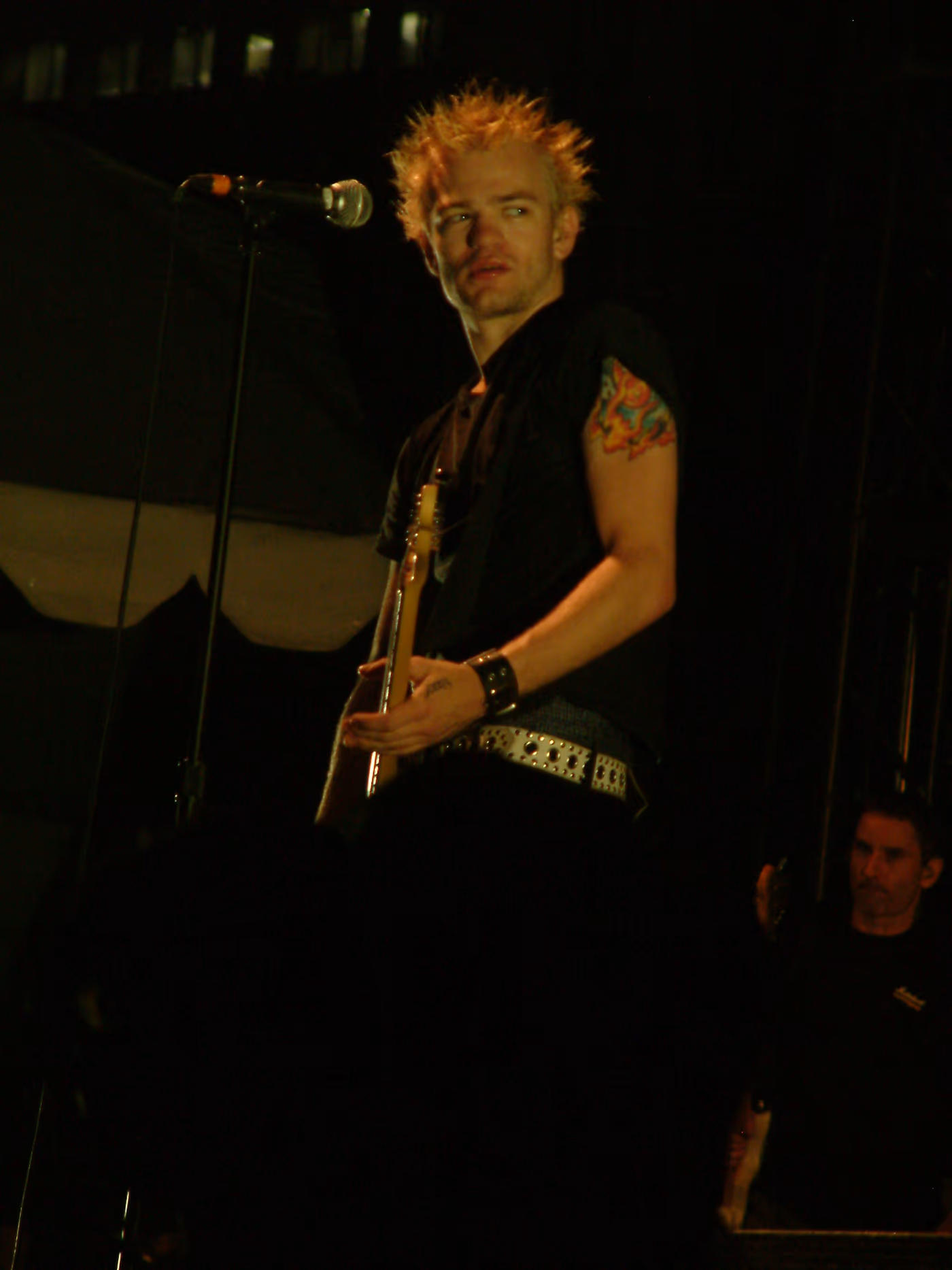 Deryck Whibley of Sum 41 on stage in 2003, with bleached spiky hair, a black sleeveless T-shirt, a flame tattoo on his upper arm and a studded belt, holding a guitar next to a microphone stand.