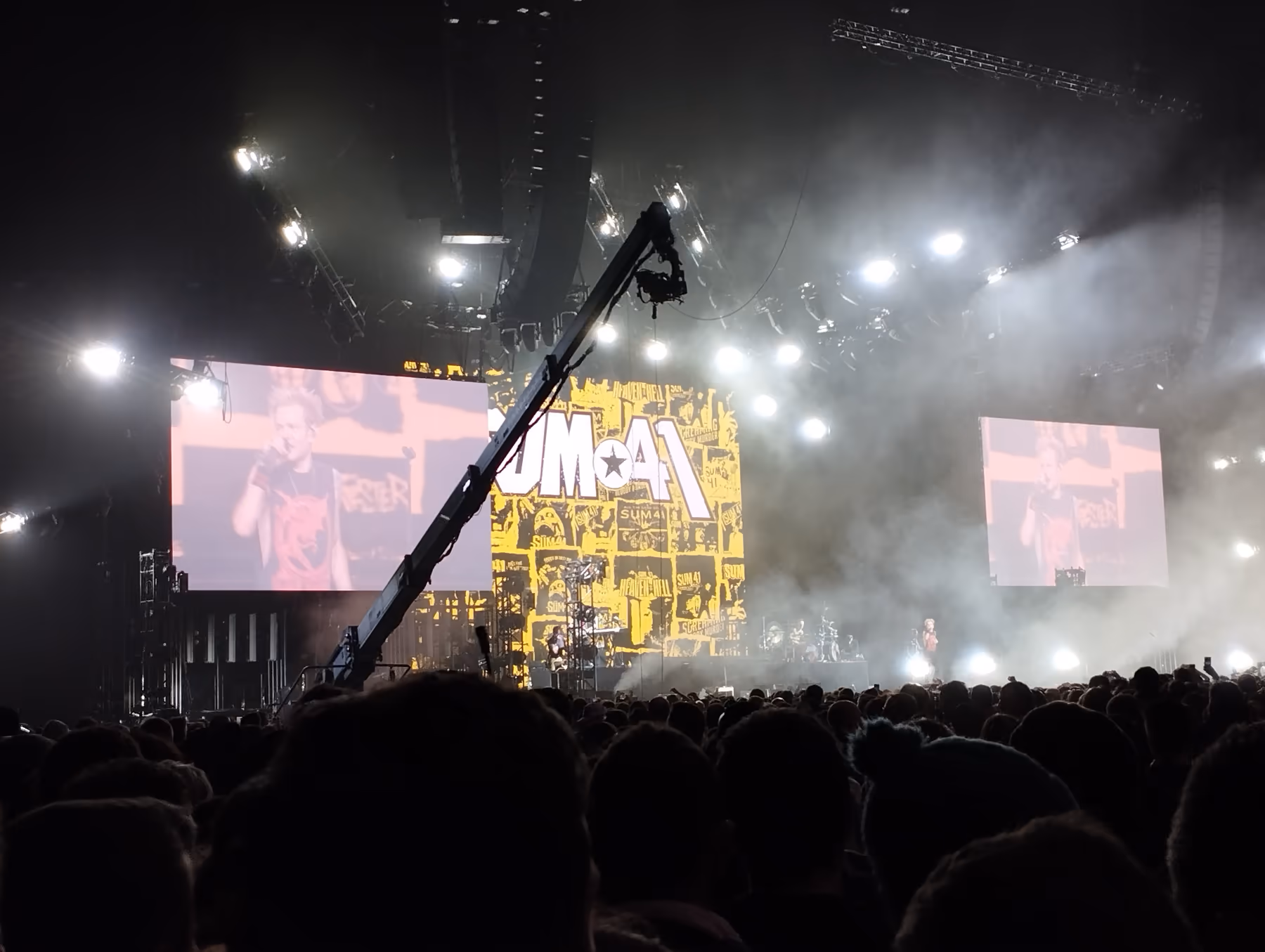 Sum 41 playing an arena show in 2024, with a large yellow stage backdrop carrying the band's logo, two video screens showing the singer, and a packed crowd silhouetted in the foreground.