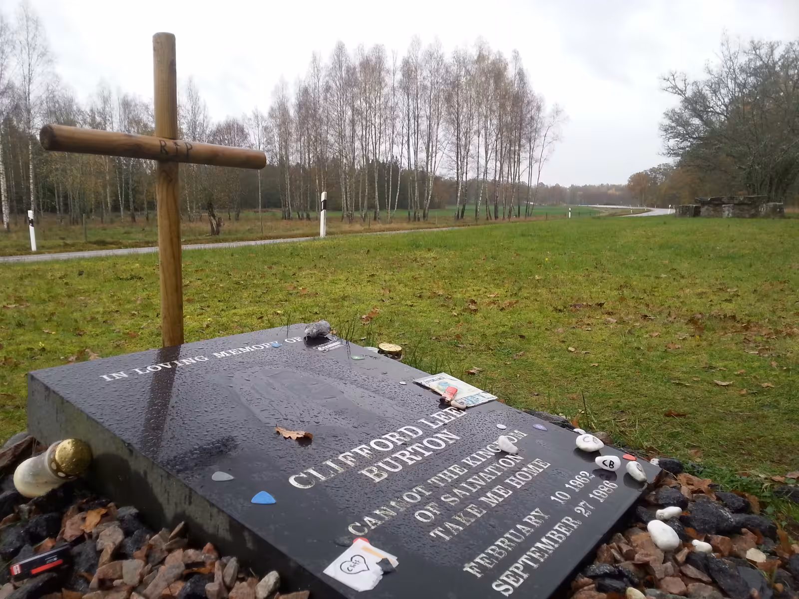 A stone memorial to Cliff Burton beside a road in the Swedish countryside, with flowers at its base.