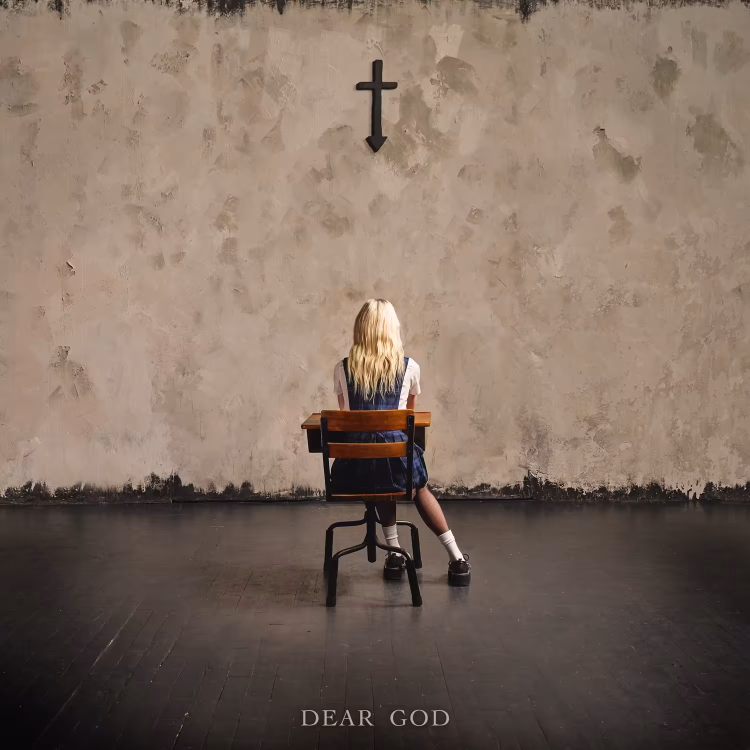 A blonde woman in a navy tartan school pinafore sits at a wooden school desk facing a bare plaster wall, with a black cross mounted above her that tapers into a downward arrow.