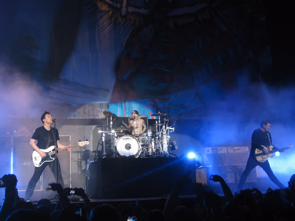 Blink-182 performing live in an arena under blue stage lighting: Mark Hoppus on bass at the left at a microphone, Travis Barker shirtless on a raised drum riser at centre, and Tom DeLonge on guitar at the right, with a crowd of raised hands in the foreground