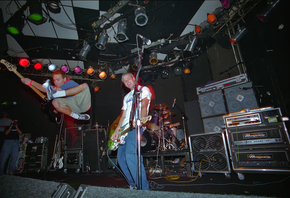 Blink-182 performing in a small club in 1996: bassist Mark Hoppus leaping mid-air with his bass while Tom DeLonge sings into the microphone with a guitar and drummer Scott Raynor plays behind them under coloured stage lights