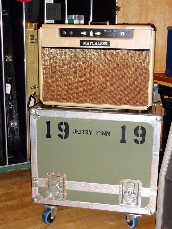 A Matchless guitar amplifier sitting on top of a green road case stencilled with the name JERRY FINN in black lettering, surrounded by flight cases in a recording studio