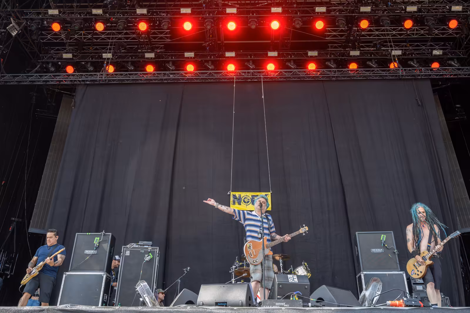 NOFX playing on a festival stage in daylight, with Fat Mike centre on bass, a guitarist either side, Mesa cabinets and red truss lighting overhead.