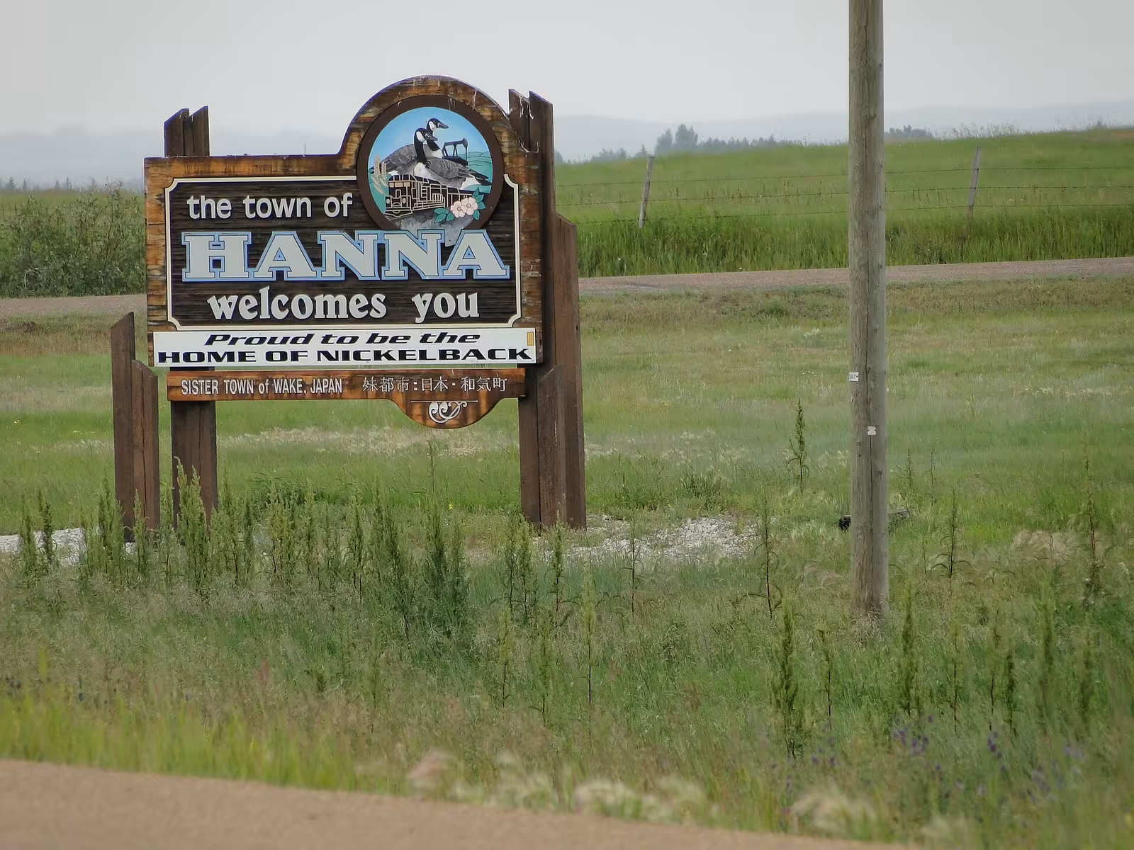 Wooden roadside sign reading 'the town of Hanna welcomes you', with a white panel beneath saying 'Proud to be the home of Nickelback' and a smaller board naming Wake, Japan as a sister town, standing in green prairie grass.
