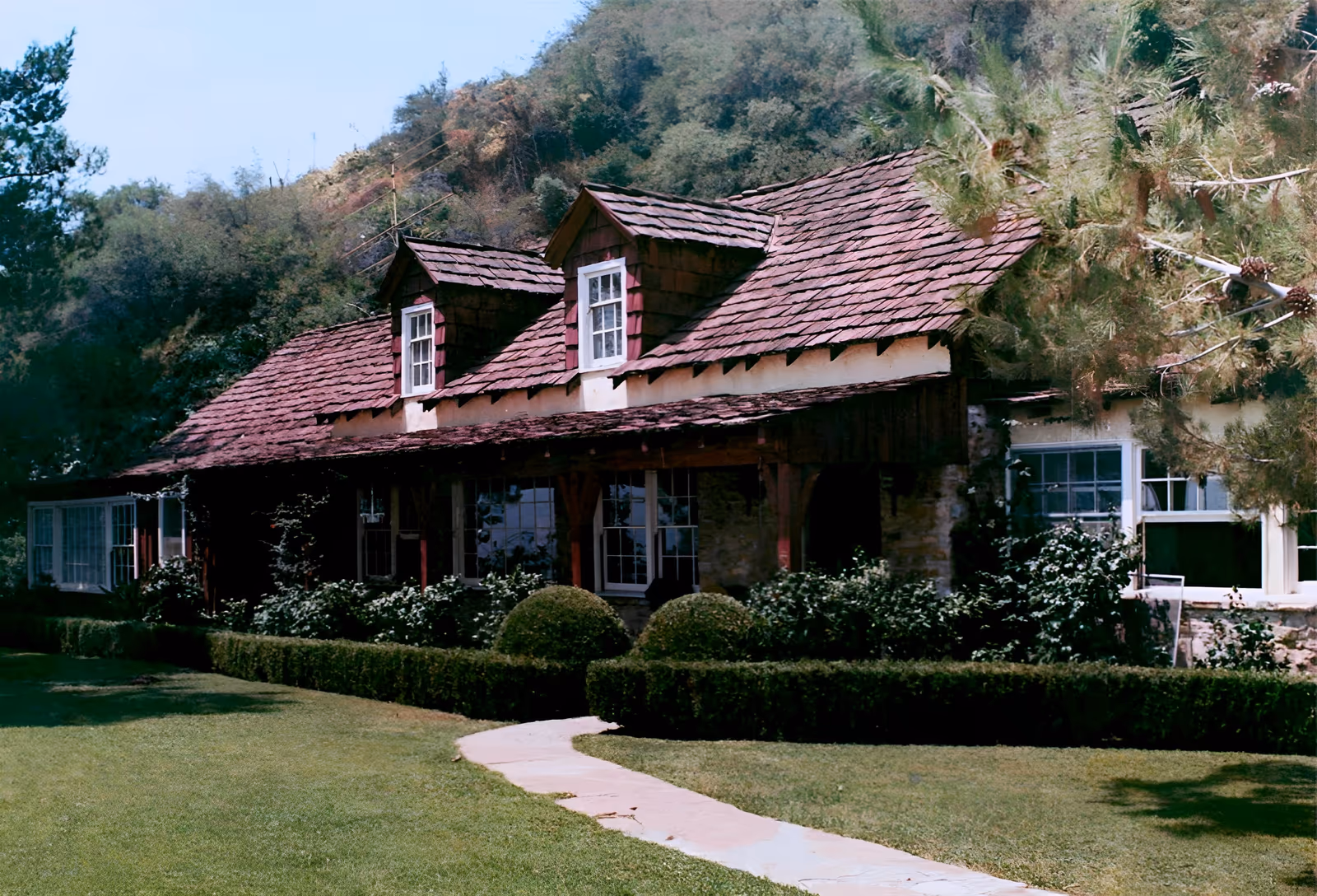 Daylight exterior of a single-storey ranch-style house with a steep shingled roof, two dormer windows, white-framed windows and clipped hedges beside a curving stone path, set against a wooded hillside.