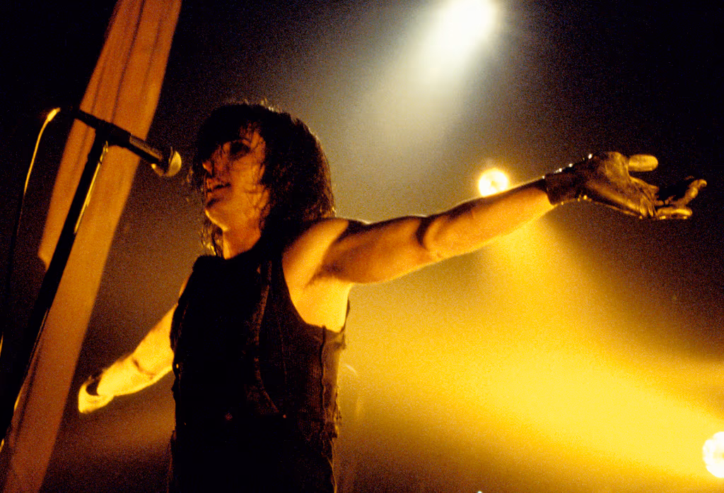 Trent Reznor on stage with wet, tangled dark hair and a sleeveless top, his right arm stretched out wearing a torn fishnet glove, singing into a microphone on a stand in front of a hanging curtain, lit from behind by yellow spotlights.