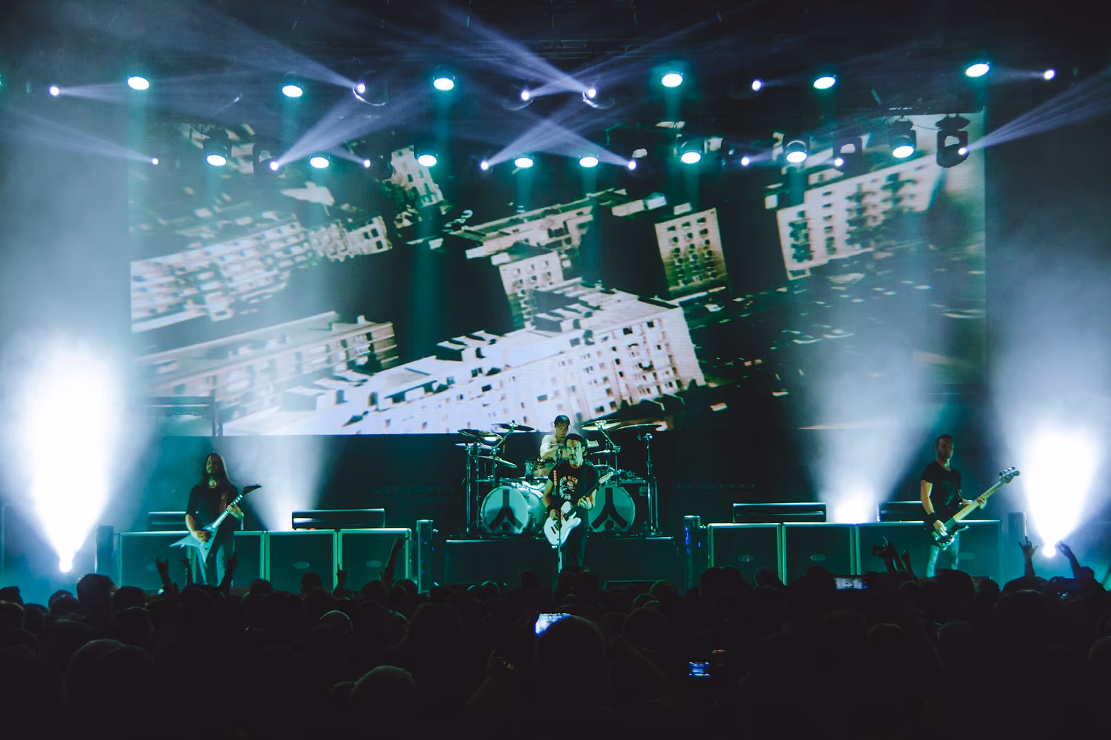 Gojira on stage in teal light in Toronto in October 2016, with a large projection screen of fragmented white shapes behind the drum riser and a crowd silhouetted in the foreground.